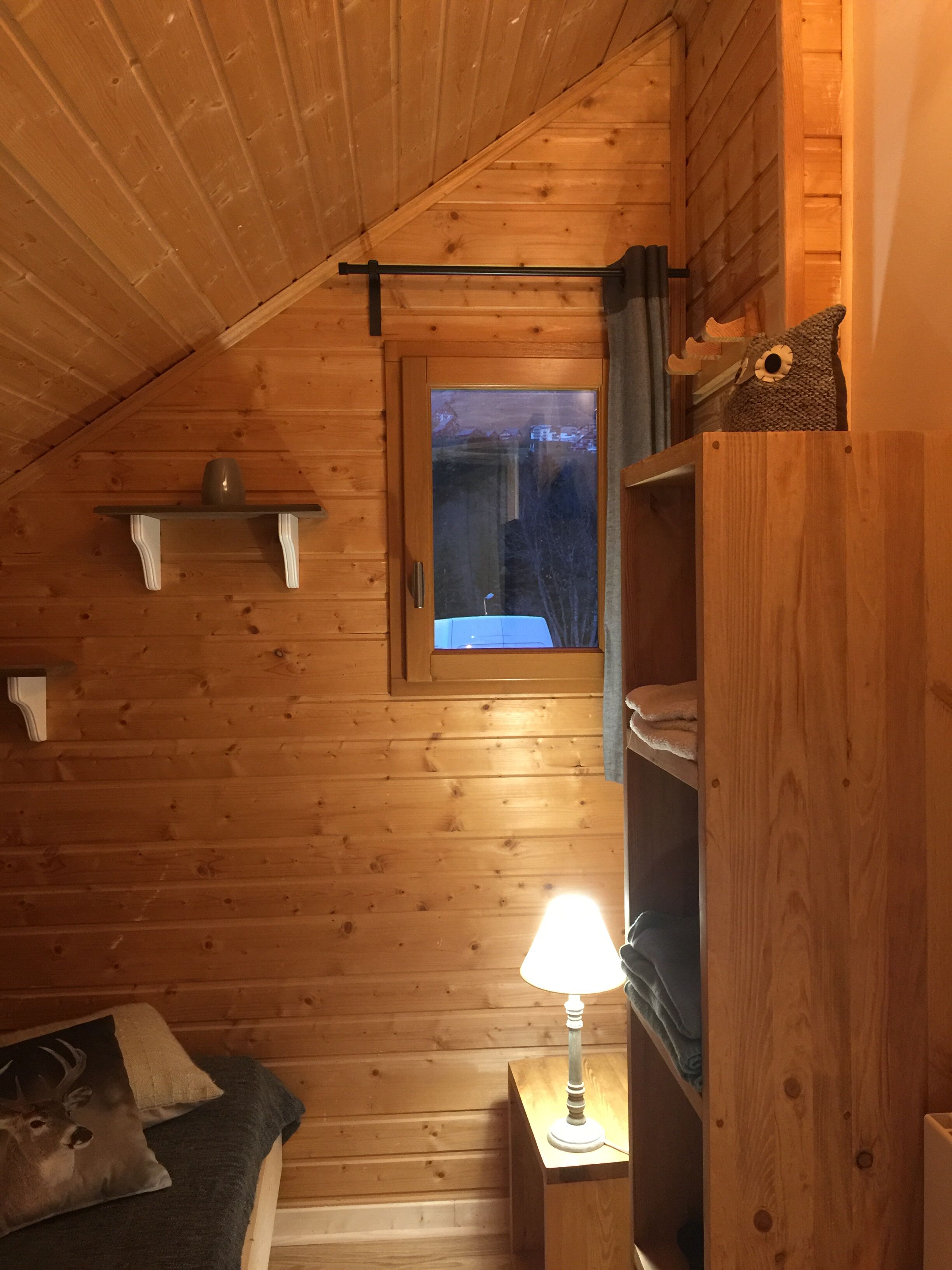A cozy cabin room with wooden walls, a window with a view, a bed with a deer pillow, a lamp, and a shelf with a stuffed animal.