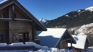 Una cabaña de madera con techo inclinado, rodeada de terreno cubierto de nieve y montañas al fondo.