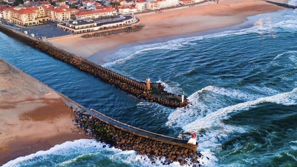 An aerial view of a coastal town with a prominent pier extending into the sea, a beach, and a dense cluster of buildings in the background.