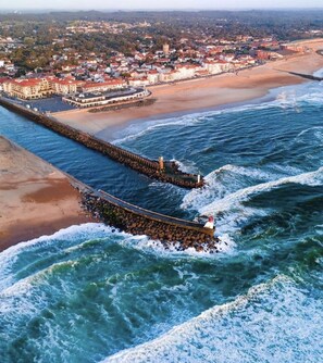 Vista aérea de un pueblo costero con un muelle destacado que se adentra en el mar, una playa y un grupo denso de edificios al fondo.