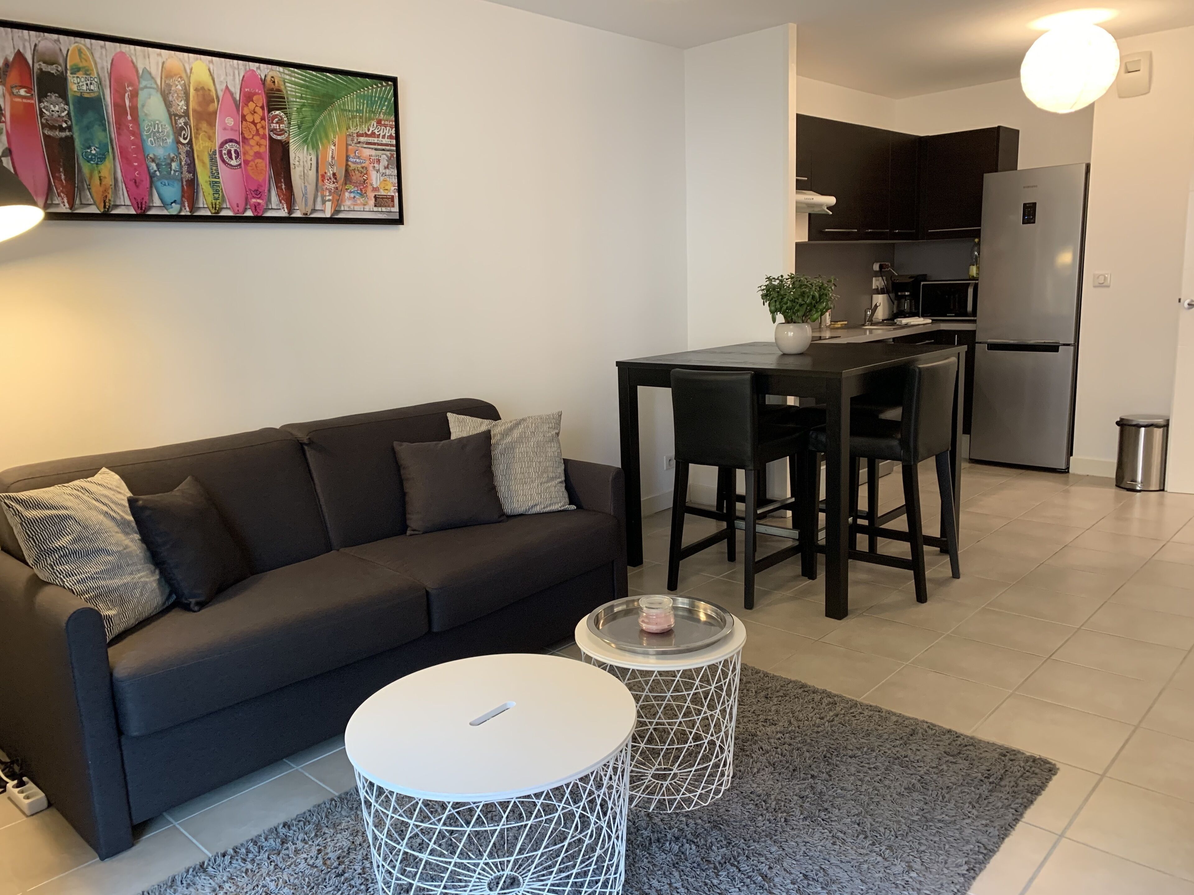 En moderne stue med en mørk sofa, et lille rundt bord og et køkkenområde med et køleskab og en mikrobølgeovn.