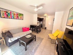 A living room with a sofa, a coffee table, a yellow armchair, a TV, and a dining area with a kitchen in the background.