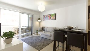 A modern living room with a sofa, dining table, and chairs. There is a sliding glass door leading to a balcony.