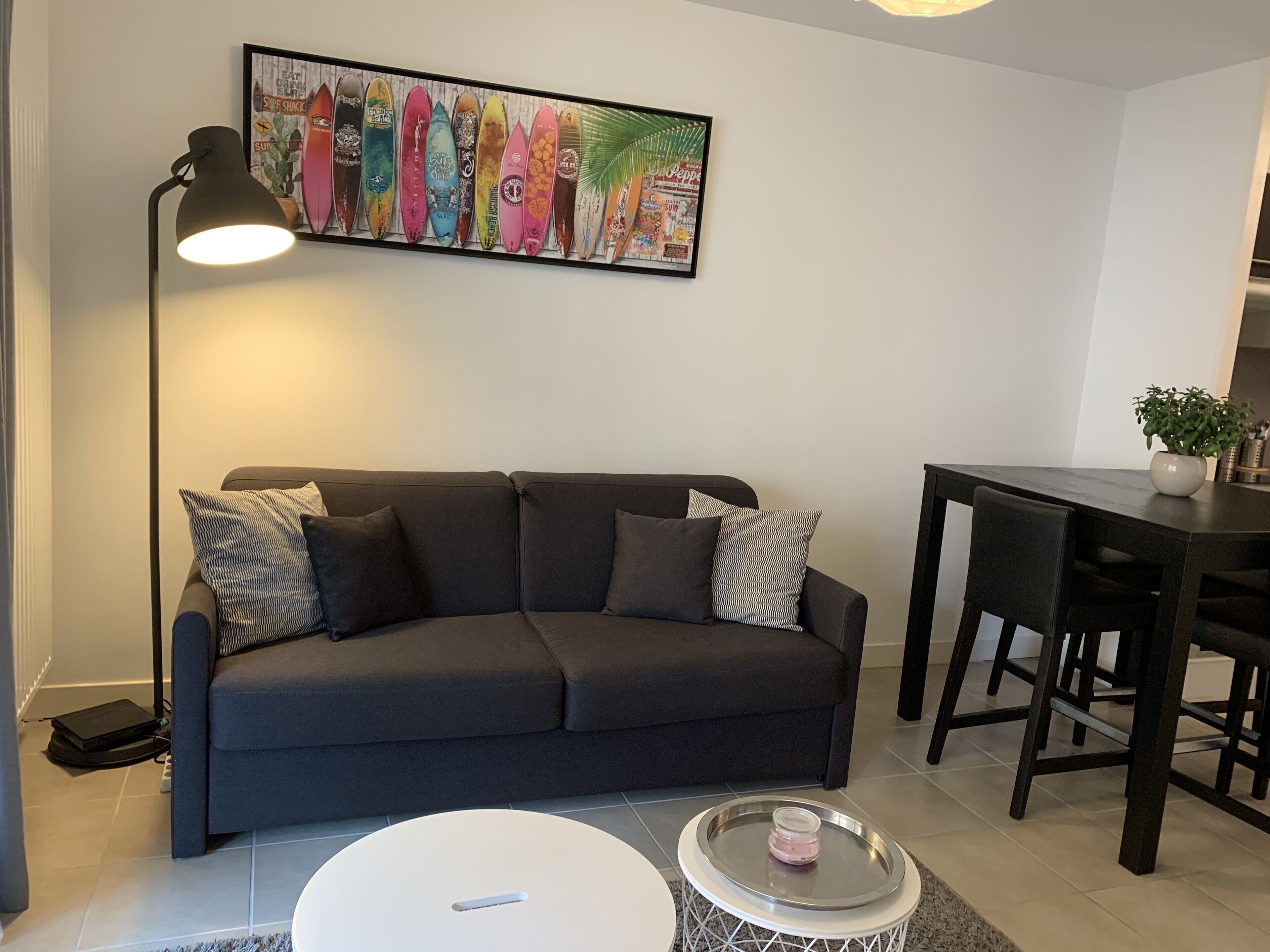 Una sala moderna con un sofá gris oscuro, una mesa de centro blanca y una mesa de comedor con sillas. También se observa una lámpara de pie y una obra de arte con forma de tabla de surf enmarcada.
