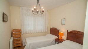 Une chambre avec deux lits simples, une table de chevet en bois avec une lampe, un meuble de rangement en osier, un radiateur et un lustre.