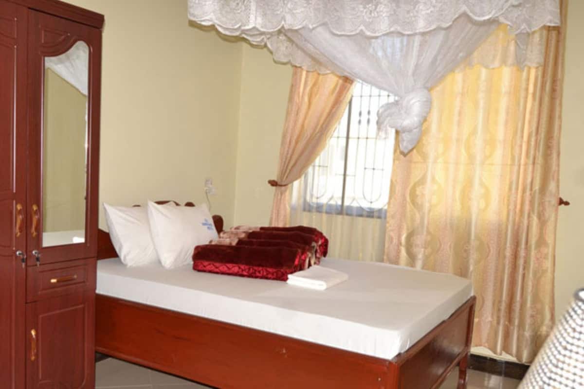 Un dormitorio con un armario de madera, una cama con ropa de cama blanca, una manta roja y una ventana con cortinas.
