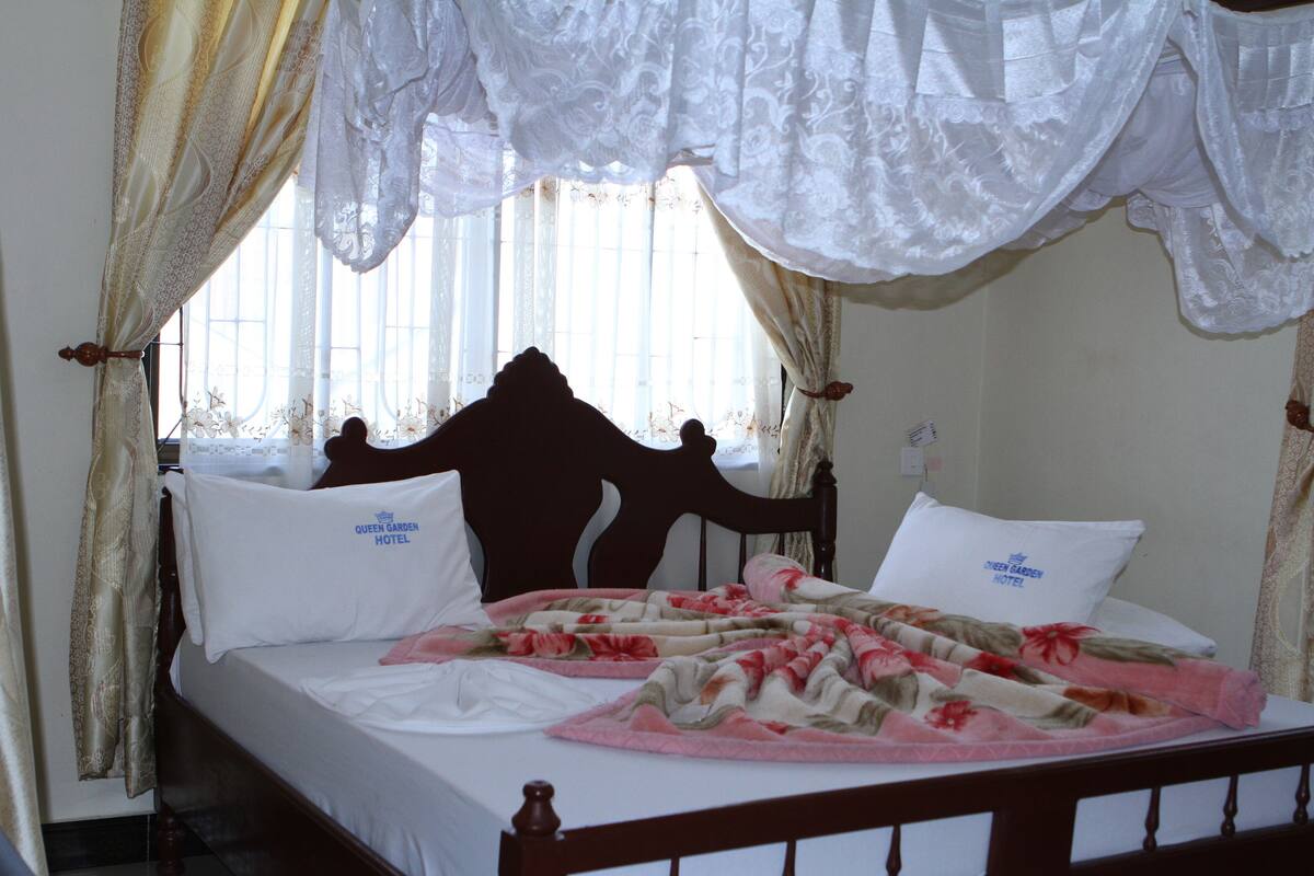 Una cama con dosel tapizada con sábanas blancas y ropa de cama floral, cabecera de madera y cortinas con encaje.