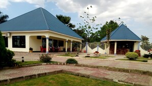 Un edificio de dos pisos con techo azul, un patio cubierto con asientos al aire libre y un jardín ajardinado con un sendero pavimentado.