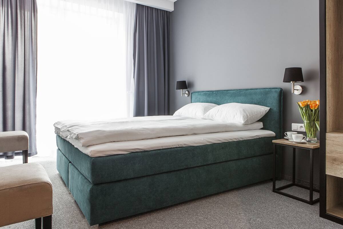 A neatly made bed with white linens, a teal upholstered headboard, and a small bedside table with a lamp and a vase of flowers.