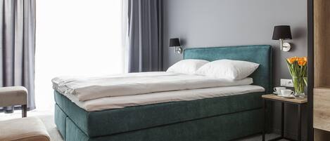 A neatly made bed with white linens, a teal upholstered headboard, and a small bedside table with a lamp and a vase of flowers.