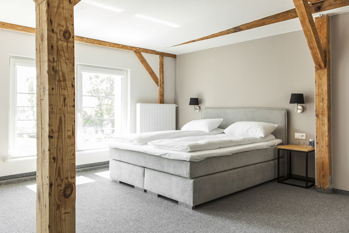 A modern bedroom with a large bed, wooden beams, a small bedside table, and a window.