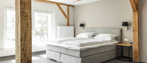 A modern bedroom with a large bed, wooden beams, a small bedside table, and a window.