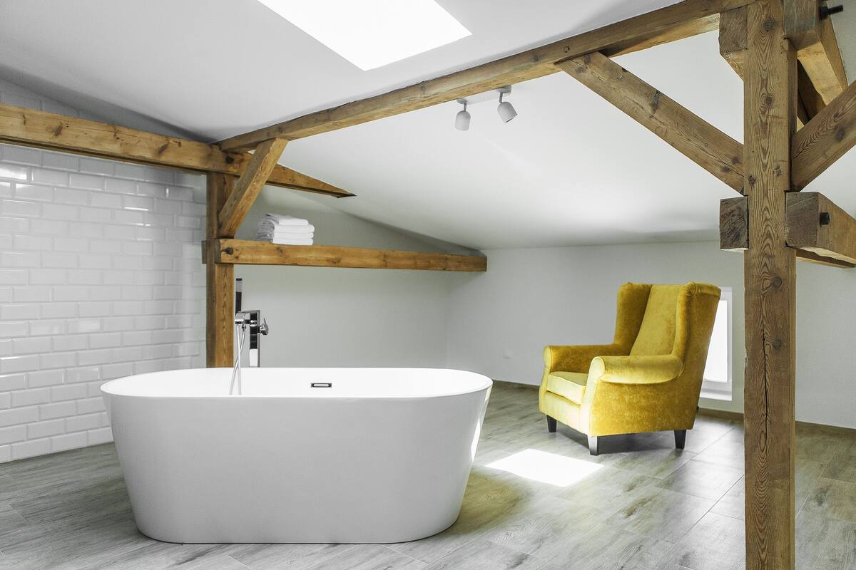 A modern bathroom with a freestanding bathtub, wooden beams, and a yellow armchair.