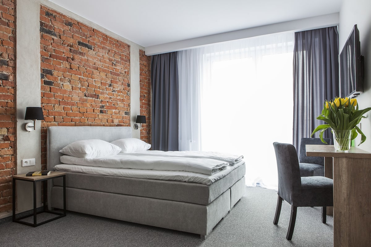 A modern hotel room with a brick wall, a bed with white linens, a desk with a chair, and a flat-screen TV.
