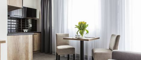 Una habitación de hotel moderna con kitchenette, un comedor con mesa y sillas, y un ventanal con cortinas translúcidas.