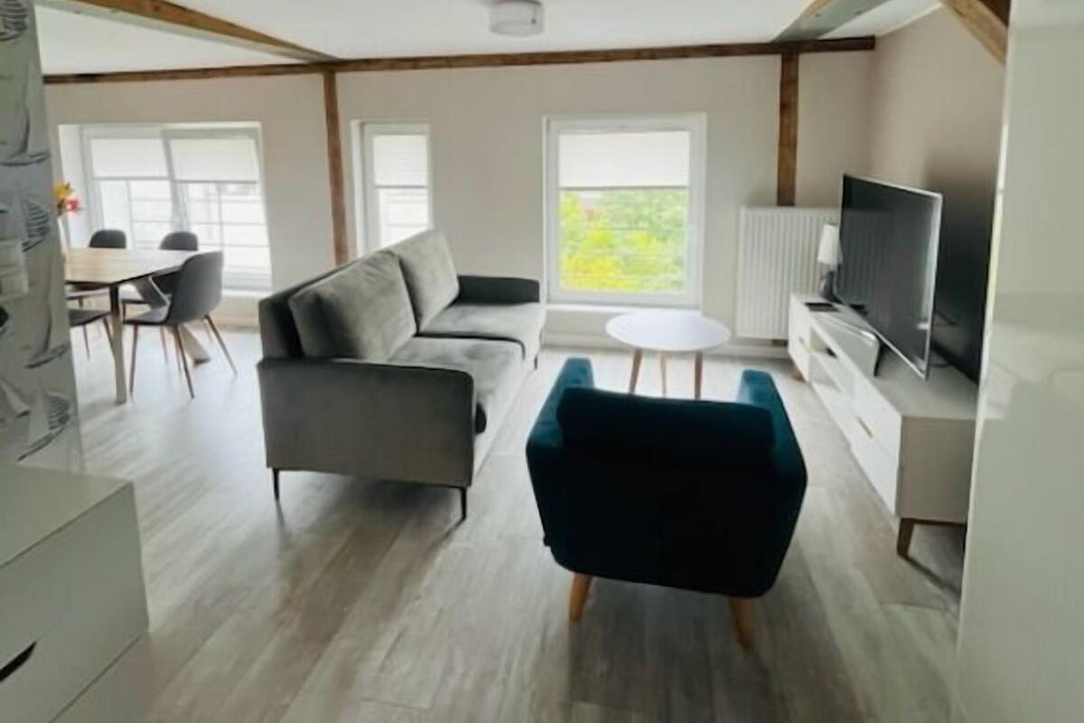 A spacious loft with a high ceiling, exposed wooden beams, a grey sofa, a teal armchair, a dining area with a table and chairs, and a TV on a white cabinet.