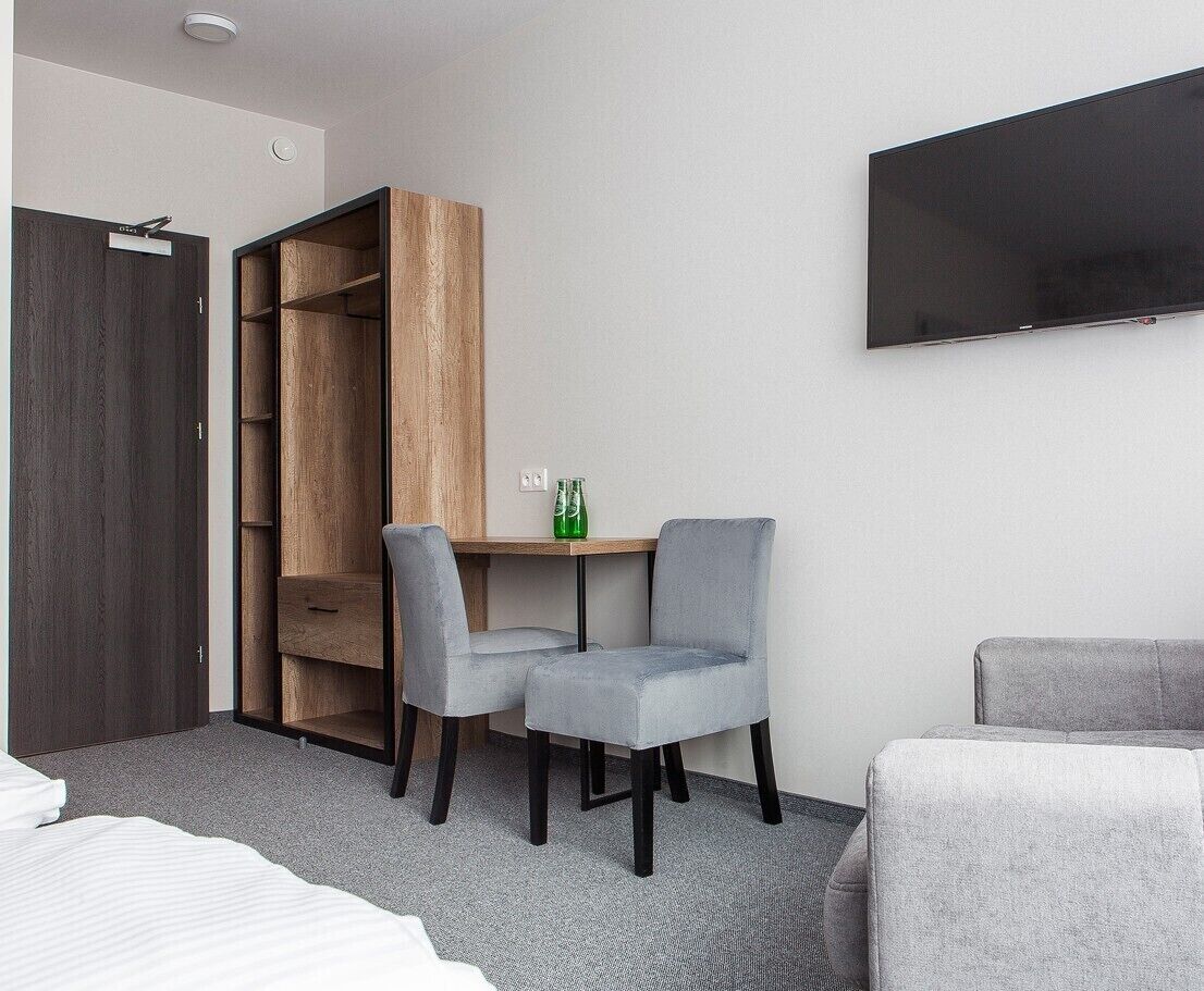 A hotel room with a bed, a grey sofa, a TV mounted on the wall, a wooden wardrobe, and a small table with two grey chairs.