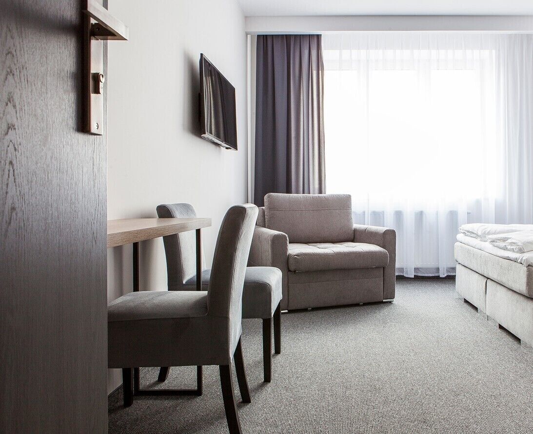 Hotellihuone, jossa on sänky, kaksi nojatuolia, pieni pöytä ja seinälle asennettu televisio.