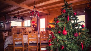 A cozy cabin interior with a wooden dining table, chairs, a Christmas tree, and a kitchen area.