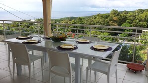 Une table à manger dressée pour quatre personnes, avec des chaises blanches, un plateau en verre et des assiettes à bordure jaune. Un vase rempli de fleurs est posé sur la table.