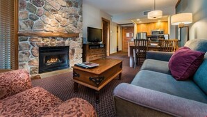 Una sala de estar con chimenea de piedra, televisor, comedor con mesa y sillas, y cocina al fondo.