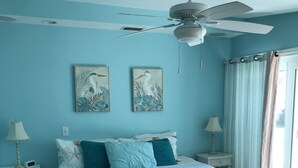Un dormitorio con cama, dos cuadros en la pared, ventilador de techo, mesita de noche con lámpara y ventana con cortinas.