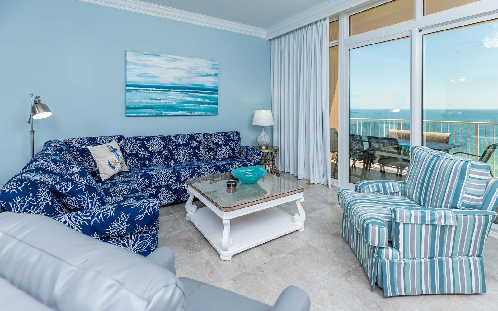 A room with a blue ocean-themed sofa, a striped armchair, and a glass coffee table.