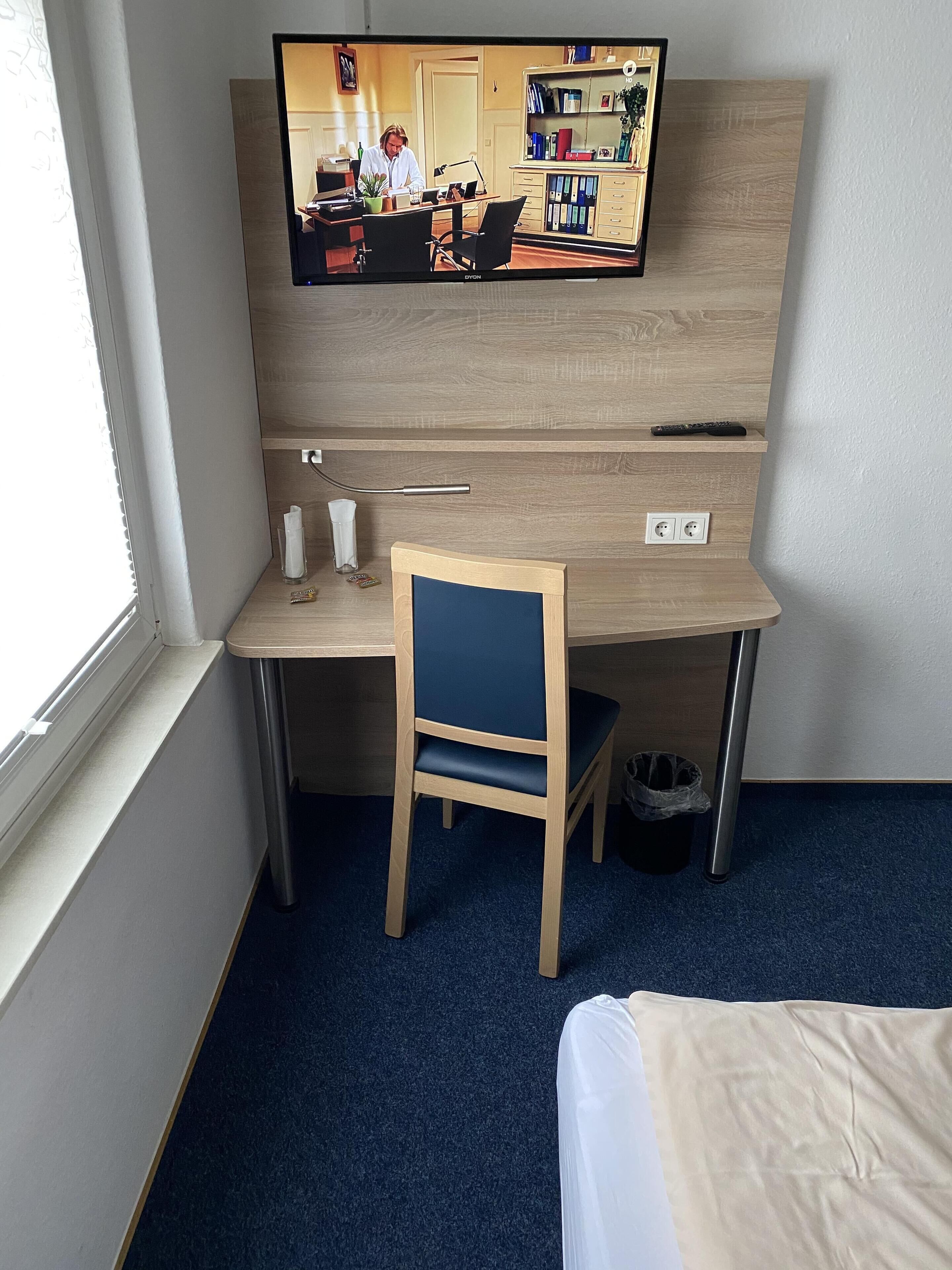 Quarto de hotel com escrivaninha, cadeira, televisão e cama.