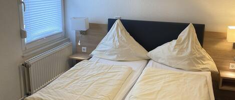Una cama doble con cabecera negra, una ventana con persianas, una mesita de noche con lámpara y un radiador.