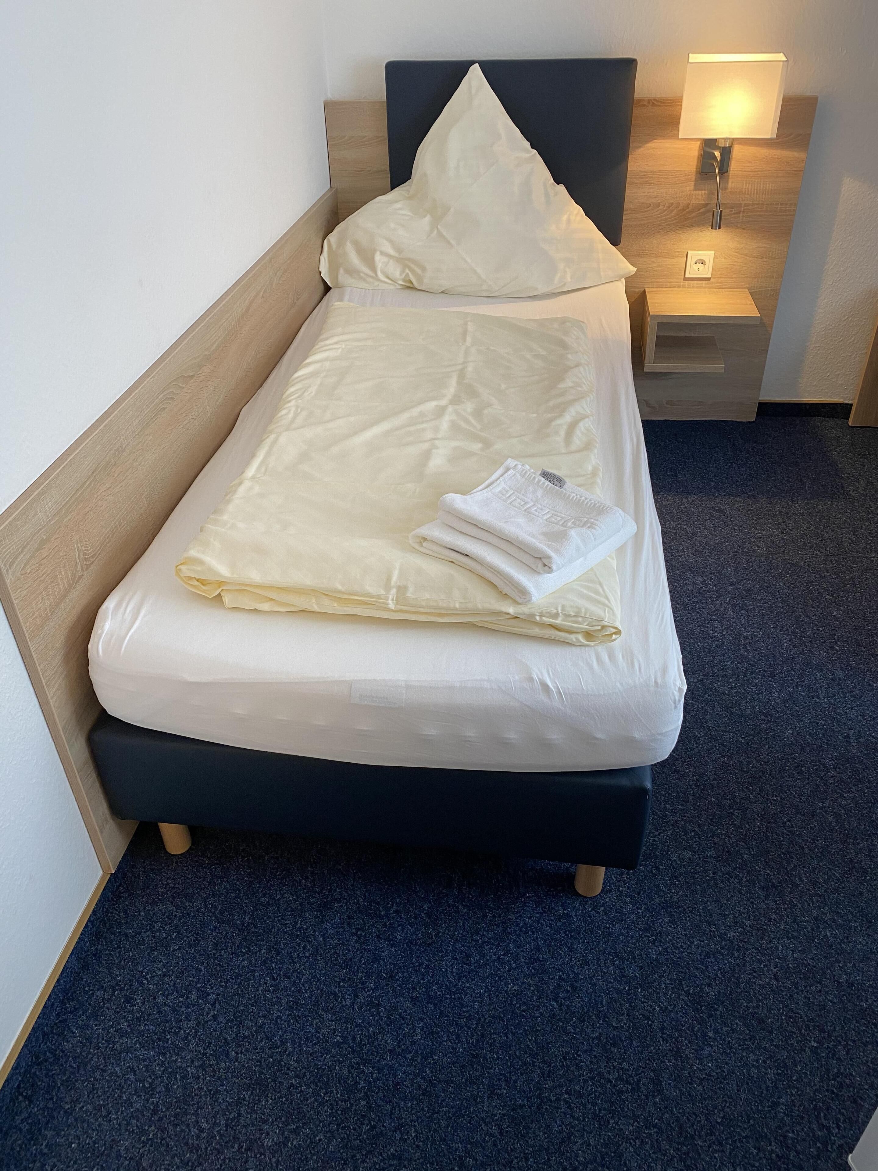 Una cama bien hecha con un colchón blanco, una manta blanca doblada y una almohada. Hay una mesita de noche con una lámpara y un enchufe. La habitación tiene un cabecero de madera y un suelo alfombrado oscuro.