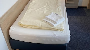Una cama bien hecha con un colchón blanco, una manta blanca doblada y una almohada. Hay una mesita de noche con una lámpara y un enchufe. La habitación tiene un cabecero de madera y un suelo alfombrado oscuro.