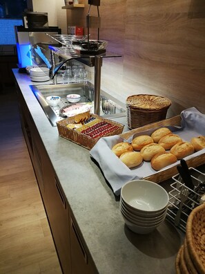 Una barra de buffet con diversos alimentos, incluyendo pan y pasteles, dispuestos sobre una mesa con cestas y fuentes.