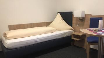 Une chambre d’hôtel de taille modeste, comprenant un lit simple, une petite table de chevet, une chaise et une table recouverte d’une nappe à carreaux.