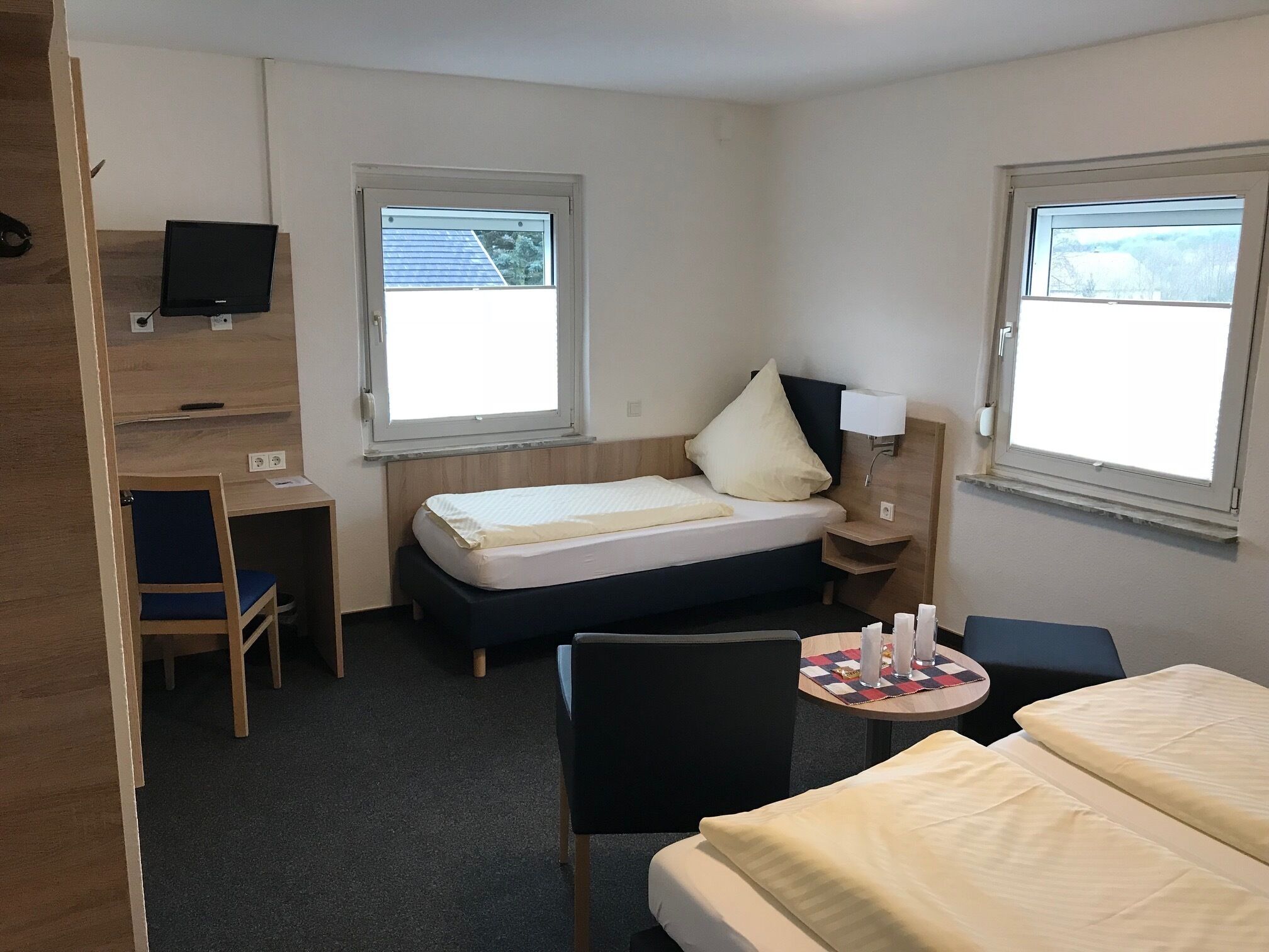A hotel room with a bed, a desk, a chair, a small table, and two windows.