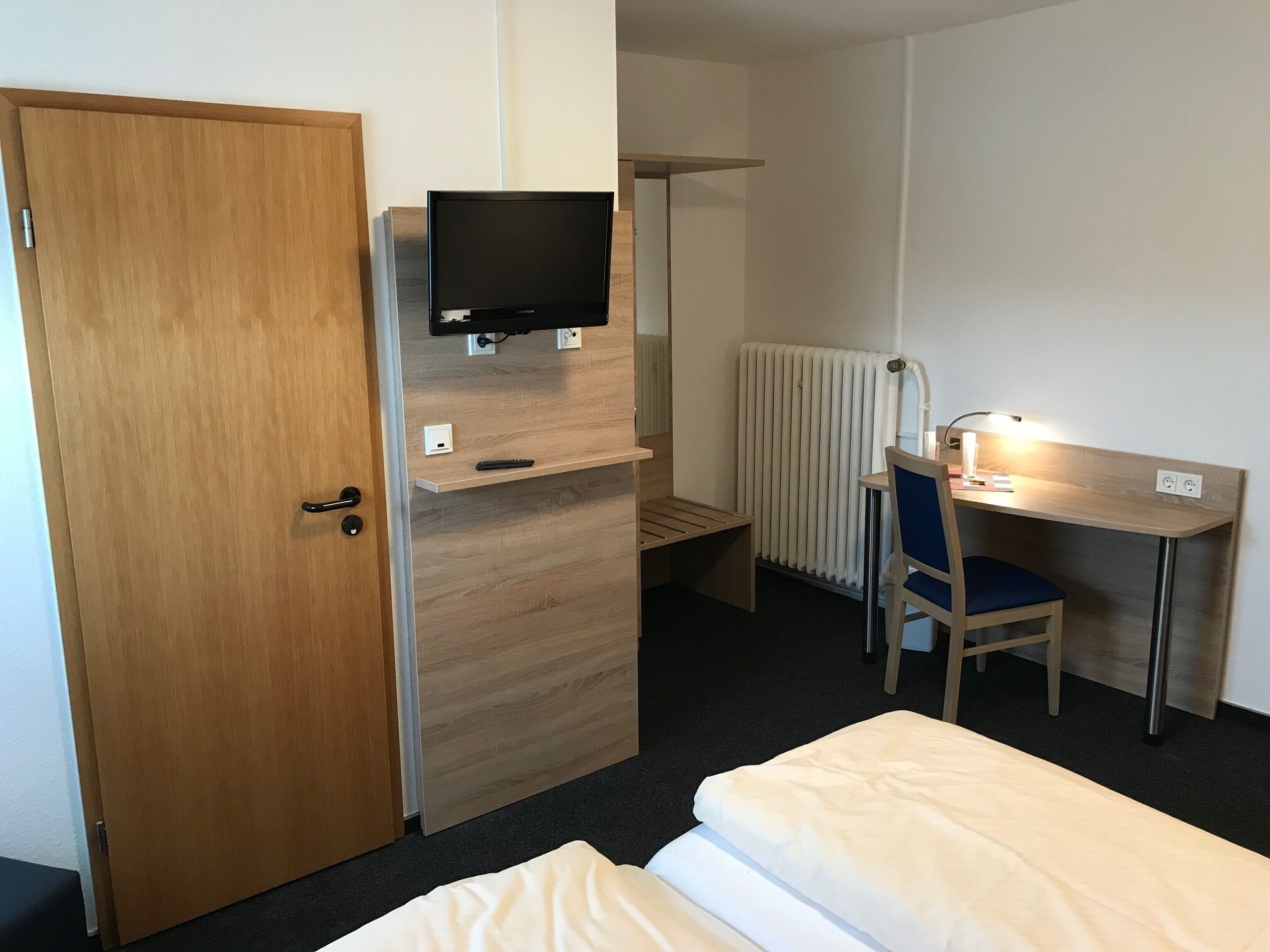 Habitación de hotel con puerta de madera, televisor montado en un estante de madera, escritorio con una silla y una cama.