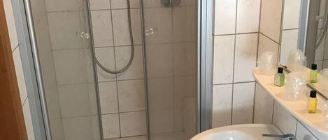 Un baño con ducha de vidrio, un lavamanos blanco con un espejo arriba y artículos de aseo en la encimera.