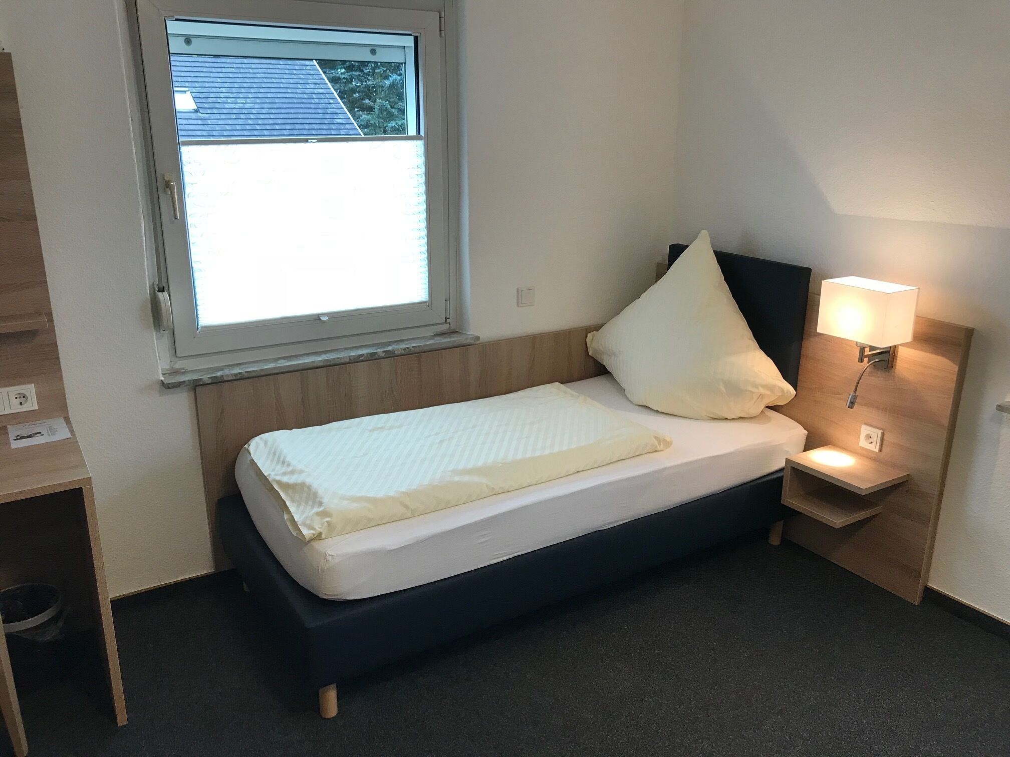 Una cama pequeña y bien hecha, con colchón blanco y una sola almohada, una mesita de noche con una lámpara, y una ventana con vistas al exterior.