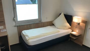 A small, neatly made bed with a white mattress and a single pillow, a bedside table with a lamp, and a window with a view of the outside.