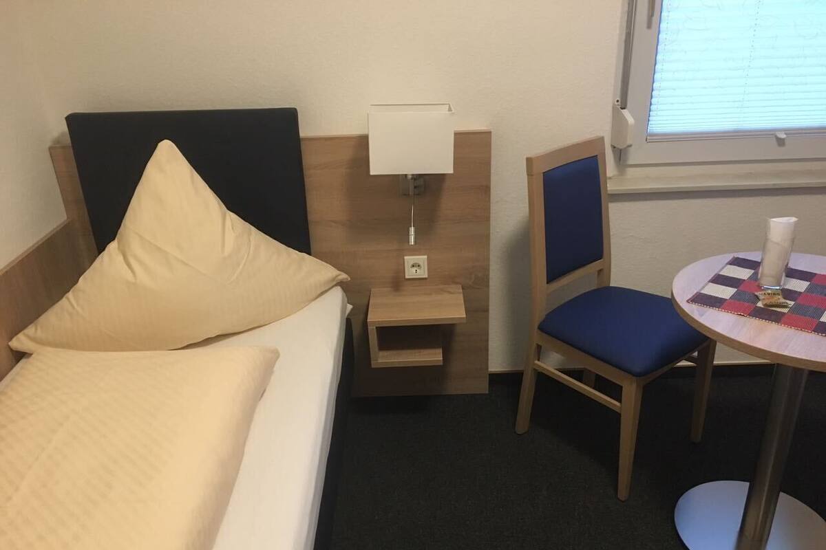 Une chambre d’hôtel avec un lit, une table de chevet avec une lampe, une chaise et une petite table recouverte d’une nappe à carreaux.