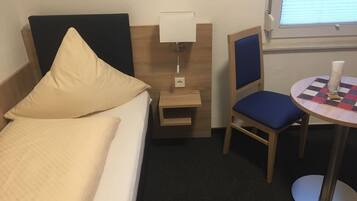 A hotel room with a bed, a nightstand with a lamp, a chair, and a small table with a checkered tablecloth.