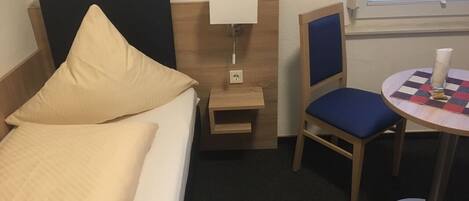 A hotel room with a bed, a nightstand with a lamp, a chair, and a small table with a checkered tablecloth.