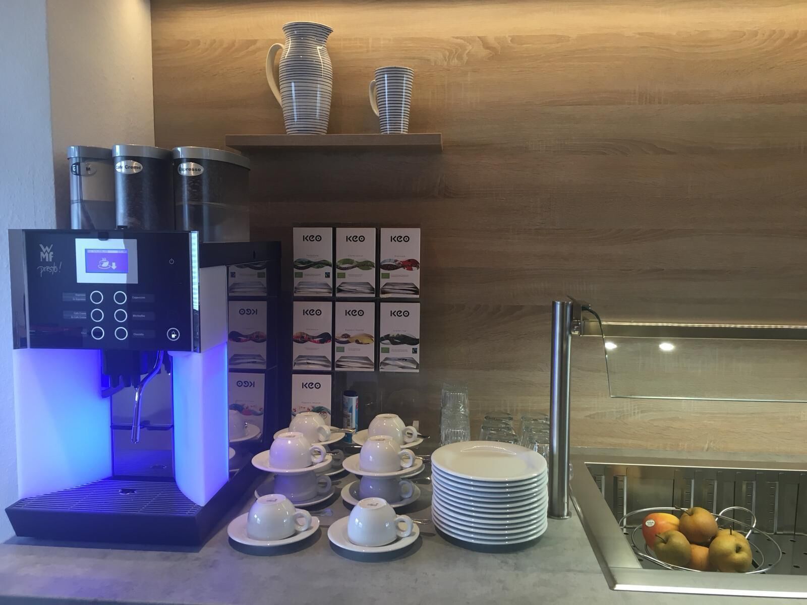 Una estación de café con una cafetera, tazas, platitos y una cesta de frutas.