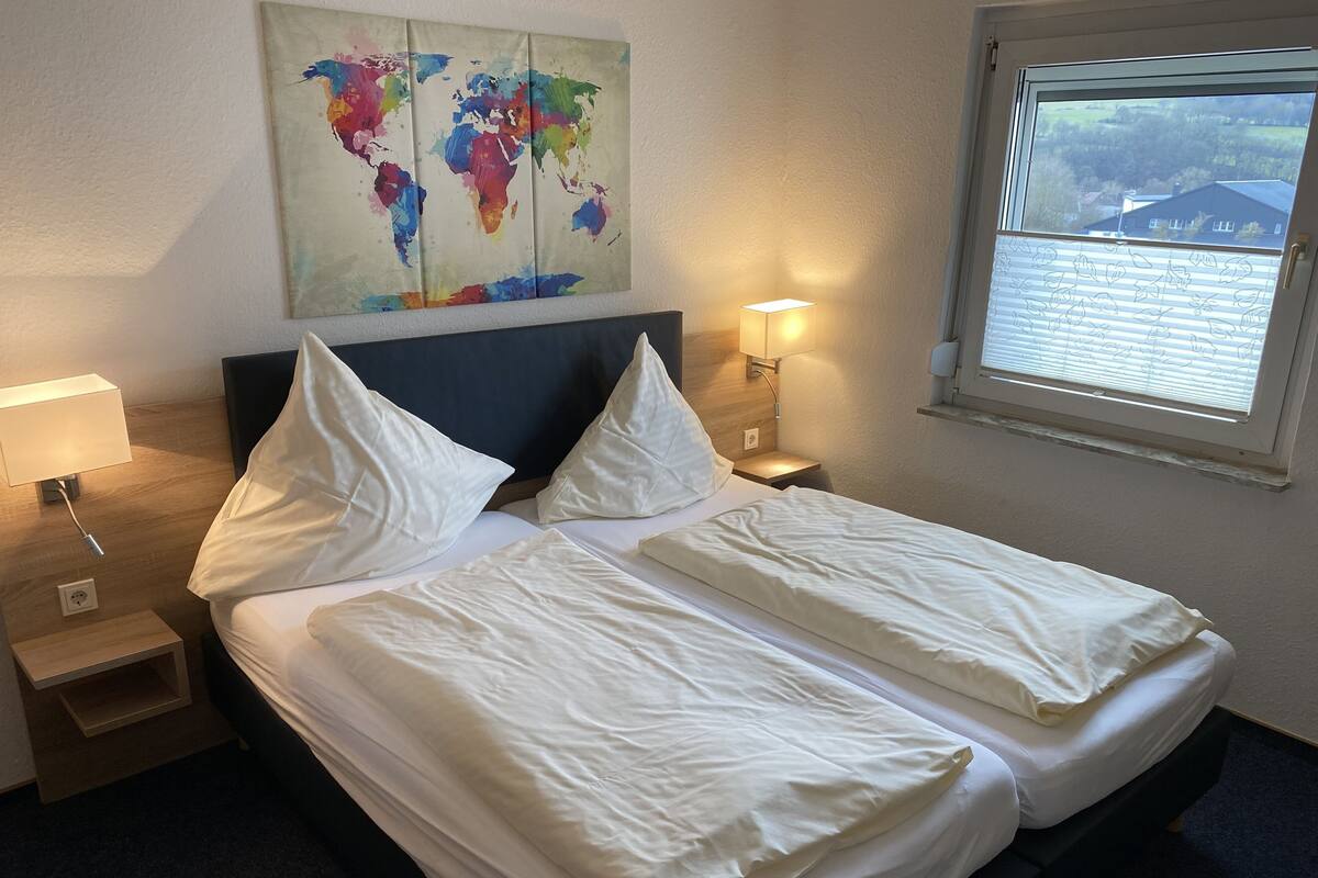 Une chambre d’hôtel avec un lit double bien fait, une œuvre d’art représentant une carte du monde accrochée au mur, une lampe de chevet et une fenêtre donnant sur la campagne.
