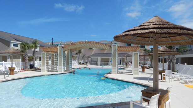 Zona de piscina amb diverses ombrel·les, gandules i una piscina blava i cristal·lina.