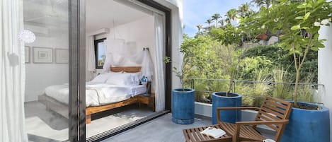 A modern bedroom with a large sliding glass door leading to a balcony with a wooden bench, a small table, and potted plants.