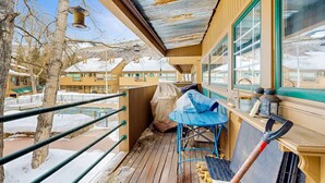 A wooden deck with a blue chair and a shovel, overlooking a snowy landscape with buildings in the background.