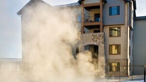 A spa pool with steam rising from the water, surrounded by a building with multiple balconies and a stone facade. - Equipped Suite | Sauna + Spa Access (Park City)