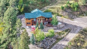 A two-story log cabin with a steeply pitched roof, surrounded by a forested area with a dirt road leading to it. - Adventures-Historic Deadwood- Trails- ATV- Skiing-Trolly-Private Acreage (Deadwood)
