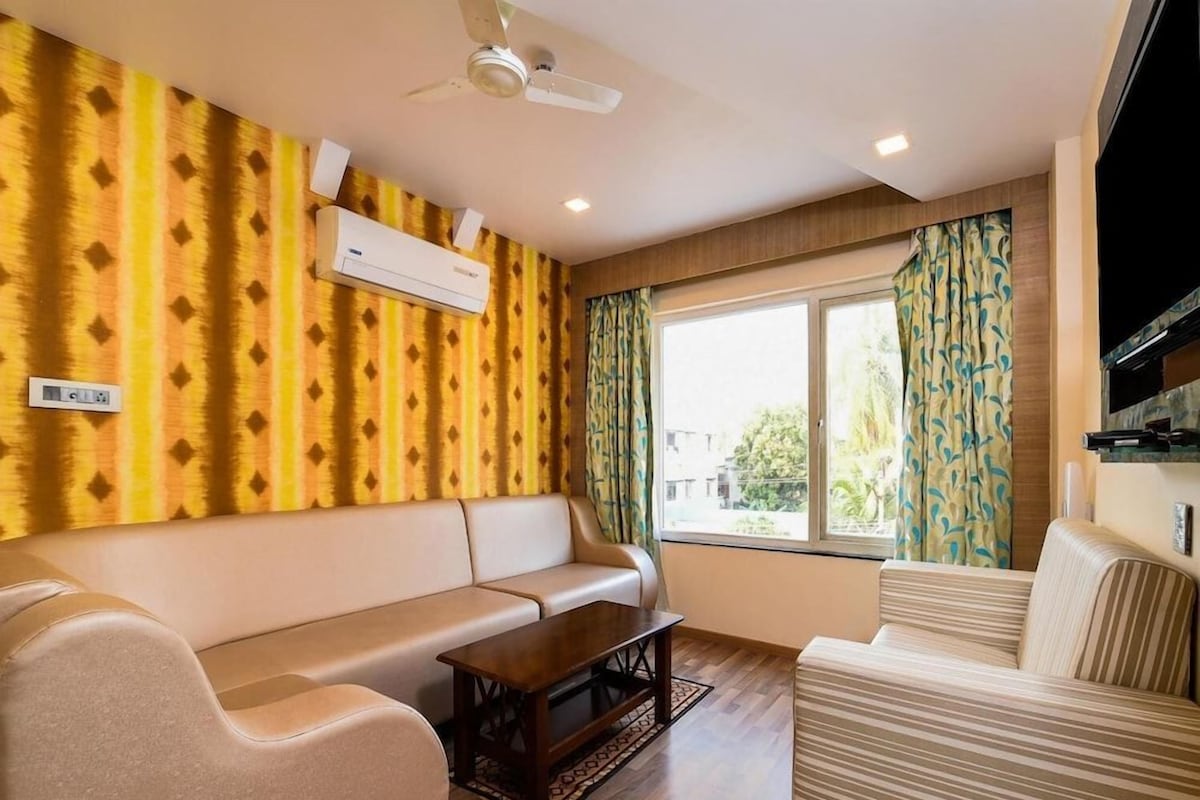 A living room with a sofa, a coffee table, and a television. There is a window with curtains, an air conditioning unit, and a ceiling fan.