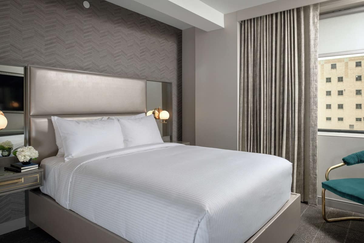 A modern hotel room with a large bed, a bedside table with a lamp, a mirror, and a view of buildings through the window.